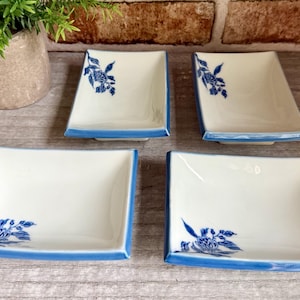 May include: A set of four rectangular ceramic dishes with a cream-colored base and a blue painted floral motif in one corner. Each dish features a blue rim and a slightly curved shape, providing a consistent look for serving small food items.