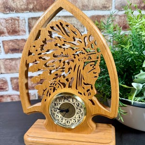 Könnte beinhalten: Eine Holzuhr mit einem Design eines indianischen Häuptlings. Das Zifferblatt der Uhr ist rund mit römischen Ziffern und einer goldfarbenen Lünette. Der hellbraune Holzrahmen hat einen dekorativen Ausschnitt eines indianischen Häuptlings.