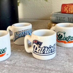 以下が含まれることがあります： NFLチームのロゴが入った4つの白い陶器製ミニチュアマグカップ。2つは緑とオレンジのマイアミ・ドルフィンズのロゴ、1つはティールと黒のジャクソンビル・ジャガーズのロゴ、もう1つは青と赤のニューイングランド・ペイトリオッツのロゴが描かれています。