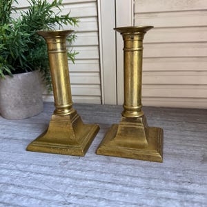 May include: A pair of matching brass candlesticks featuring a cylindrical stem with horizontal ring detailing, a flared top rim, and a square base. The metal has a polished gold-toned finish with visible signs of age and natural patina.