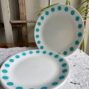 May include: A stack of white ceramic plates with a teal polka dot border. The plates are stacked on a white lace tablecloth. The plates are round and have a simple, yet elegant design.