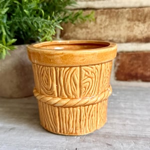 May include: A cylindrical ceramic planter with a glossy golden-yellow finish. The exterior features a molded relief pattern resembling vertical wooden planks and a horizontal rope detail encircling the middle of the vessel, creating a textured look.