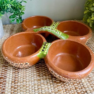 May include: A four-section terracotta serving dish featuring a natural reddish-brown clay finish. The bowls are connected by a central bridge painted with green accents and small yellow and black dots, with matching dot patterns along the outer edges.