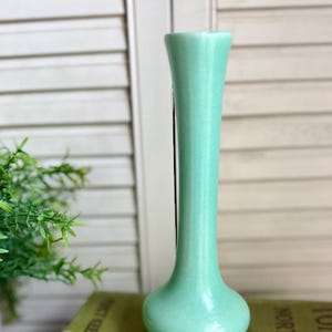 May include: A tall, slender bud vase with a smooth, matte finish in a soft mint green color. The vase features a narrow neck that flares slightly at the top opening and widens into a rounded base, creating a simple, vertical silhouette.