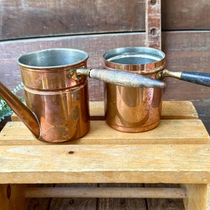 May include: Two cylindrical copper pots with metallic interiors and long, dark wooden handles. One pot features a prominent, angled copper spout extending from the side. Both vessels have a polished exterior finish and are positioned side by side.