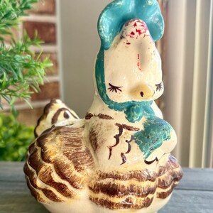 May include: A ceramic chicken figurine featuring a cream-colored body with brown painted feather details. The chicken wears a bright blue bonnet tied under its chin, has closed eyes with long eyelashes, and a small red mark on its forehead.