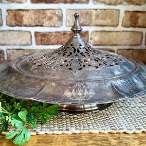 May include: A silver-toned metal serving dish with a wide, flat rim and a removable, domed lid. The lid features an intricate, openwork filigree pattern and a central finial. The base of the dish is decorated with subtle, etched floral motifs.