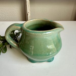Op de afbeelding: Kleine groene keramische kan met een afgeronde body, een gebogen handvat en een schenktuit. De kan heeft een glanzende afwerking en staat op drie kleine voetjes. De kleur is een gedempt, zeegroen.