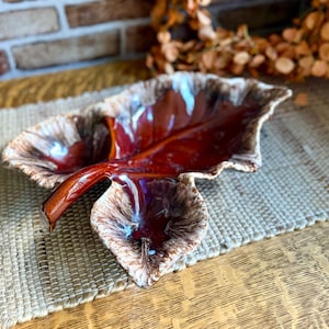 May include: A ceramic leaf-shaped dish featuring a glossy deep red and brown center glaze that transitions into a textured, cream and brown mottled edge. The dish has a prominent stem handle and a realistic, organic silhouette with raised veins.
