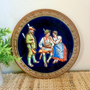 Może przedstawiać: Okrągły ozdobny talerz ceramiczny z ciemnoniebieskim środkiem przedstawiającym wypukłą scenę z trzema postaciami w tradycyjnych strojach. Mężczyzna w zieleni stoi z karabinem, a drugi gra na cytrze dla kobiety w niebiesko-czerwonej sukni.