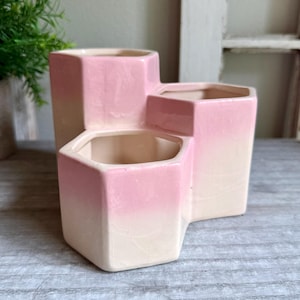 May include: A ceramic desk organizer featuring three hexagonal compartments of varying heights. The piece has a glossy finish with a gradient color scheme, transitioning from a soft cream at the base to a vibrant pink at the top, with visible crackle glaze.