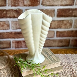 May include: A cream-colored ceramic vase with a fluted, shell-like design and a scalloped rim. The vase features a vertical ribbed texture that tapers toward a circular base, presenting a smooth, matte finish throughout its structure.