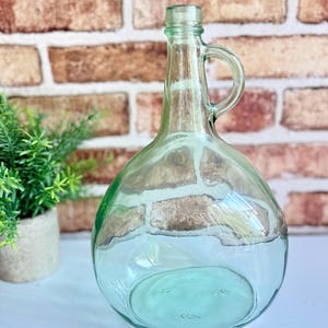 May include: A clear, light green glass bottle with a rounded body, a narrow neck, and a small integrated handle. The bottle features a thick, circular base and a simple rimmed opening at the top, showcasing a transparent, smooth glass construction.