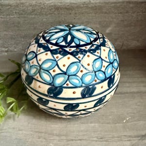 May include: A spherical ceramic container featuring a cream-colored base with intricate hand-painted patterns in shades of dark navy and light blue. The design includes floral-like motifs, geometric lines, and small brown dots across the surface.