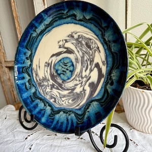 Puede incluir: Un plato de cerámica con un diseño único, con un patrón arremolinado en el centro con tonos de gris y blanco. El borde es azul oscuro con un aspecto texturizado. El plato se exhibe en un soporte de metal negro.