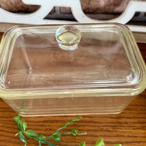 May include: A clear, rectangular glass dish with a matching lid and a small, round knob. The dish has a slightly yellow tint and is sitting on a wooden surface. The lid fits snugly, and the knob is easy to grip.