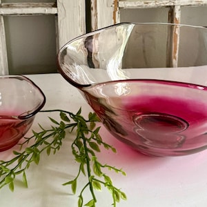 May include: Two clear glass bowls with a deep magenta hue at the base. The bowls have an organic, asymmetrical shape. A sprig of green foliage is placed between the bowls, adding a natural element to the composition.