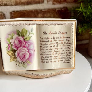 Op de afbeelding: Een crèmekleurige, boekvormige keramische plantenbak met gouden rand. De linkerpagina bevat een illustratie van roze rozen met groene bladeren. De rechterpagina toont de tekst van het Onze Vader in gouden schrift met een gouden afwerking.