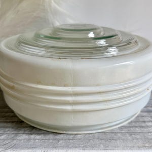 May include: A round, white milk glass light fixture shade featuring a clear glass center with concentric circular ridges. The opaque white body has three horizontal embossed rings around the exterior circumference, showing minor surface wear.