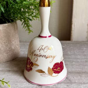 Può includere: Una campana in ceramica bianca con punta del manico dorata, un sottile bordo rosa e la scritta 40th Anniversary in corsivo dorato. La campana è decorata con illustrazioni floreali rosse e dorate di fiori e foglie attorno alla base.
