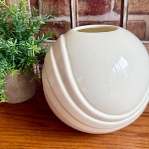 May include: Cream-colored, spherical vase with a wide opening. The vase features a series of curved, ridged details around its surface. A small potted plant with green foliage is visible to the left of the vase. The vase is sitting on a wooden surface.