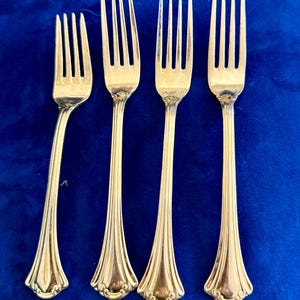 May include: A set of four gold-toned metal forks arranged vertically. Each fork features a slender handle with a flared base, decorative embossed detailing at the bottom, and four tines. The metal has a polished, reflective finish throughout.