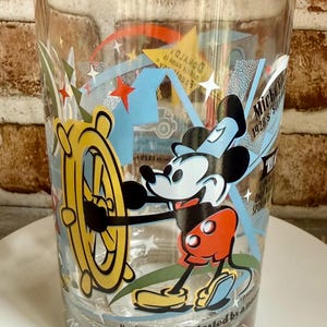 May include: A clear glass tumbler featuring a colorful graphic of Mickey Mouse at a ship's wheel. The design includes yellow stars, blue geometric shapes, and text that reads "Mickey Mouse 1928's Steamboat Willie" and "...it was all started by a mouse."