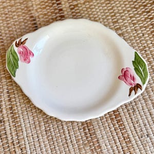 May include: A cream-colored ceramic serving dish with a scalloped edge and integrated handles on both sides. The dish features hand-painted floral motifs on each handle, consisting of pink tulip-like blossoms with green leaves and brown stems.