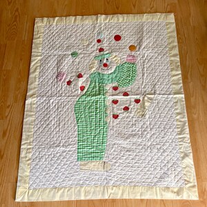 May include: A rectangular quilted blanket featuring a central applique of a clown juggling red strawberries. The clown wears a green and white checkered outfit with a pale yellow border. The background is white with a diamond-stitched pattern.
