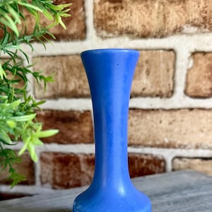 May include: A solid blue bud vase with a smooth, matte finish. The vase features a slender, cylindrical neck that gently flares outward at the top opening and widens slightly at the base, creating a simple, vertical, and symmetrical silhouette.