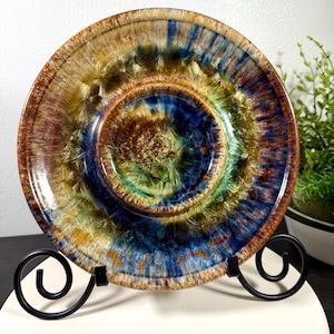 Puede incluir: Un plato decorativo de cerámica con un esmalte único. El plato presenta un diseño concéntrico con colores que van del marrón y el dorado al azul y el verde. El plato se exhibe sobre un soporte de metal negro, mostrando sus detalles artísticos.
