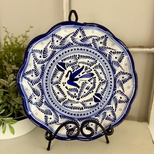 May include: A round ceramic plate with a scalloped edge, featuring a white base and intricate cobalt blue hand-painted patterns. The center displays a stylized bird surrounded by repeating feather-like motifs, all framed by concentric circular borders.