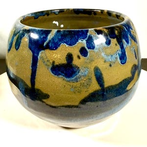 May include: A handcrafted ceramic bowl with a rounded shape. The bowl features a tan base with dark blue glaze dripping down the sides. The interior is a darker shade of tan. The bowl is suitable for decorative or functional use.