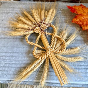 Peut inclure: Une gerbe de blé décorative composée de tiges dorées disposées en éventail, agrémentée de deux petites roses en paille tressée et de boucles en paille finement entrelacées. Le faisceau est maintenu par une fine ficelle, créant un ornement texturé.