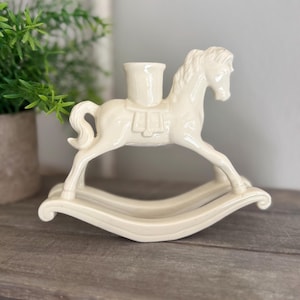 May include: A cream-colored ceramic rocking horse candle holder featuring a smooth, glossy finish. The horse is depicted in a standing position on curved rockers, with a cylindrical candle cup centered on its back, showcasing a sculpted mane and tail.