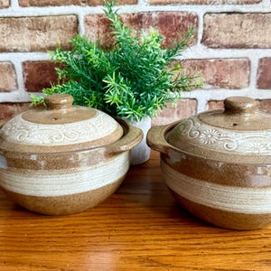 May include: Two ceramic lidded bowls with a rustic design. The bowls are brown with a cream-colored band around the middle and floral detailing on the lids. The bowls have handles on the sides and are sitting on a wooden surface.