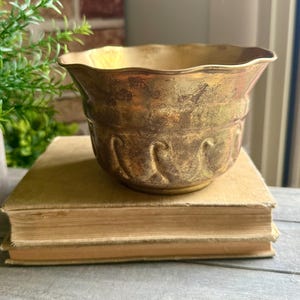 Puede incluir: Un cuenco de metal dorado con un borde festoneado y una superficie texturizada que presenta patrones en relieve en forma de onda en el exterior. El cuenco tiene un acabado envejecido y desgastado con variaciones visibles de color en el metal.
