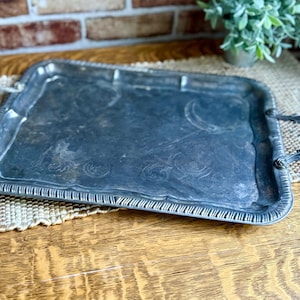 May include: A rectangular silver-toned metal serving tray featuring ornate, curved handles on each side. The tray has a raised, textured border with vertical ridges and a central surface decorated with faint, etched floral patterns and scrollwork.
