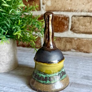 May include: A ceramic hand bell featuring a glossy brown handle and a multi-colored body. The bell has a dark brown top section, a vibrant yellow middle band, and a textured lower section with mottled green and brown glaze patterns.