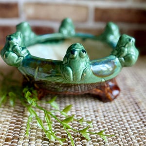 May include: A round ceramic bowl featuring a glossy green glaze with five sculpted frog figures perched along the rim. The exterior base is finished in a dark brown tone, and the interior of the bowl has a smooth, light cream-colored surface.