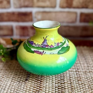 May include: A ceramic vase with a rounded body and a short neck, featuring a vibrant yellow to green gradient finish. The surface is decorated with a hand-painted scene of a Dutch landscape, including a windmill, small houses, and a canal.