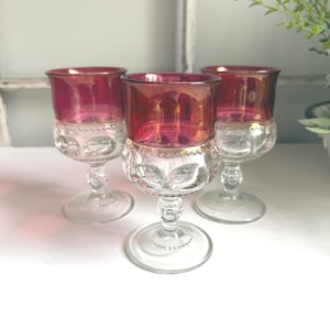May include: Three vintage wine glasses with ruby red bowls and clear, patterned bases. The glasses have a classic design with a rounded bowl and a delicate stem. The ruby red color adds a touch of elegance to the glassware.