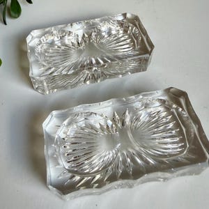 May include: Two clear, rectangular glass dishes with a decorative, sunburst-like pattern. The dishes are made of thick glass and have a faceted edge. The dishes are likely used for serving or display.