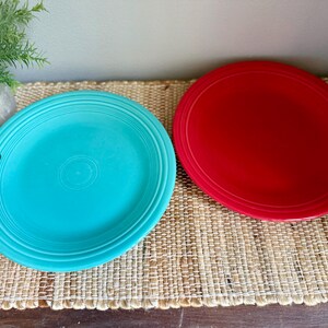 May include: Two round ceramic dinner plates, one in a bright turquoise blue and the other in a vibrant red. Both plates feature a series of concentric circular ridges embossed along the outer rim, providing a textured border to the smooth center.