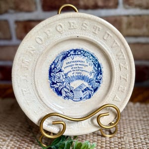 May include: A cream-colored ceramic plate featuring an embossed alphabet border and a central blue transferware design. The text reads: Drunkenness revenges the madness of one hour, with the sad repentance of many. A Holy Bible is depicted below.
