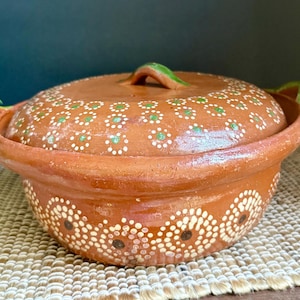May include: A round terracotta cooking pot with a matching lid, featuring hand-painted white and green floral dot patterns. The pot has two side handles and a central lid knob, all accented with green glaze, showcasing a natural clay finish.
