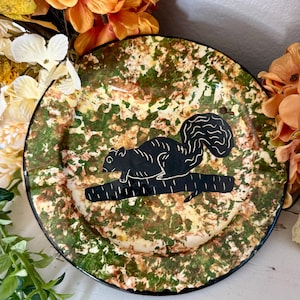 May include: A round plate featuring a mottled green and tan camouflage-like pattern across its surface. Centered on the plate is a solid black silhouette graphic of a squirrel perched on a horizontal tree branch, detailed with white line accents.