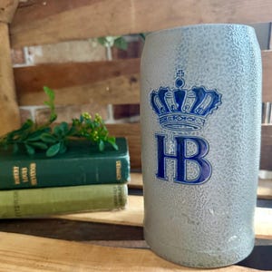 May include: A gray beer stein with a blue crown and the letters "HB". The stein is cylindrical and has a textured surface. The stein is sitting on a wooden surface.