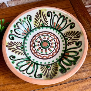 May include: A round, ceramic plate with a cream-colored interior and a peach-colored rim. The plate features a central circular design with brown, green, and orange patterns. The outer edge has a repeating floral design in green, brown, and yellow.
