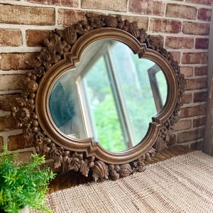 May include: A wall mirror with a quatrefoil-shaped glass pane and a brown frame featuring intricate, carved floral and fruit motifs. The frame displays detailed relief work of leaves, blossoms, and rounded fruit shapes surrounding the central glass.